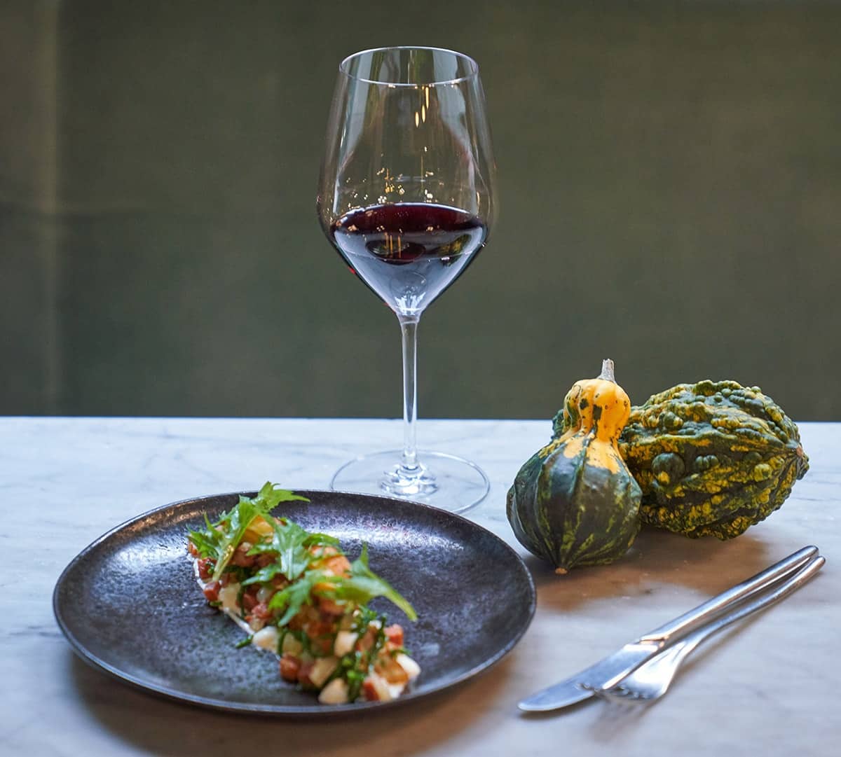 A glass of red wine shown with a plate of food and a couple gourds.